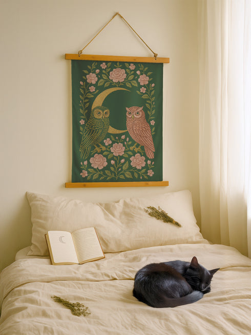 26x36 inch vertical wall tapestry printed on silky polyester, featuring a pair of embroidered-style owls perched on a golden crescent moon, surrounded by pink vintage-style florals and greenery on a forest green background. Includes stained wood slats and twine for easy hanging. A romantic, nature-inspired design for woodland and cottagecore decor lovers.
